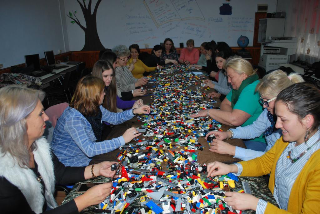 Masă lungă plină de piese LEGO, cu profesori lucrând de o parte și de alta a ei.